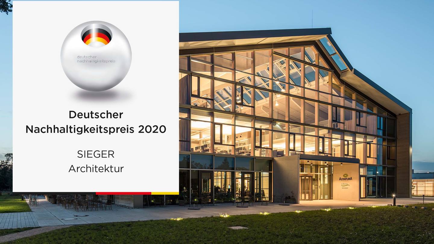 Der Alnatura Campus in Darmstadt ist zu sehen. Er ist von innen beleuchtet, da es bereits dämmert. Links auf dem Bild ist das Logo des "Deutscher Nachhaltigkeitspreis 2020" abgebildet mit dem Zusatz "Sieger Architektur"